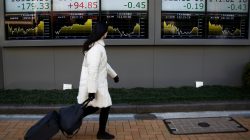 woman walks past electronic board with stock market numbers on it